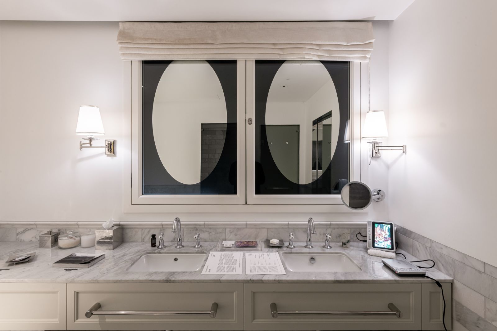 Symmetrical view of a marble double vanity with documentation papers and a small screen displaying fire. A large window with an oval reflection hangs above.