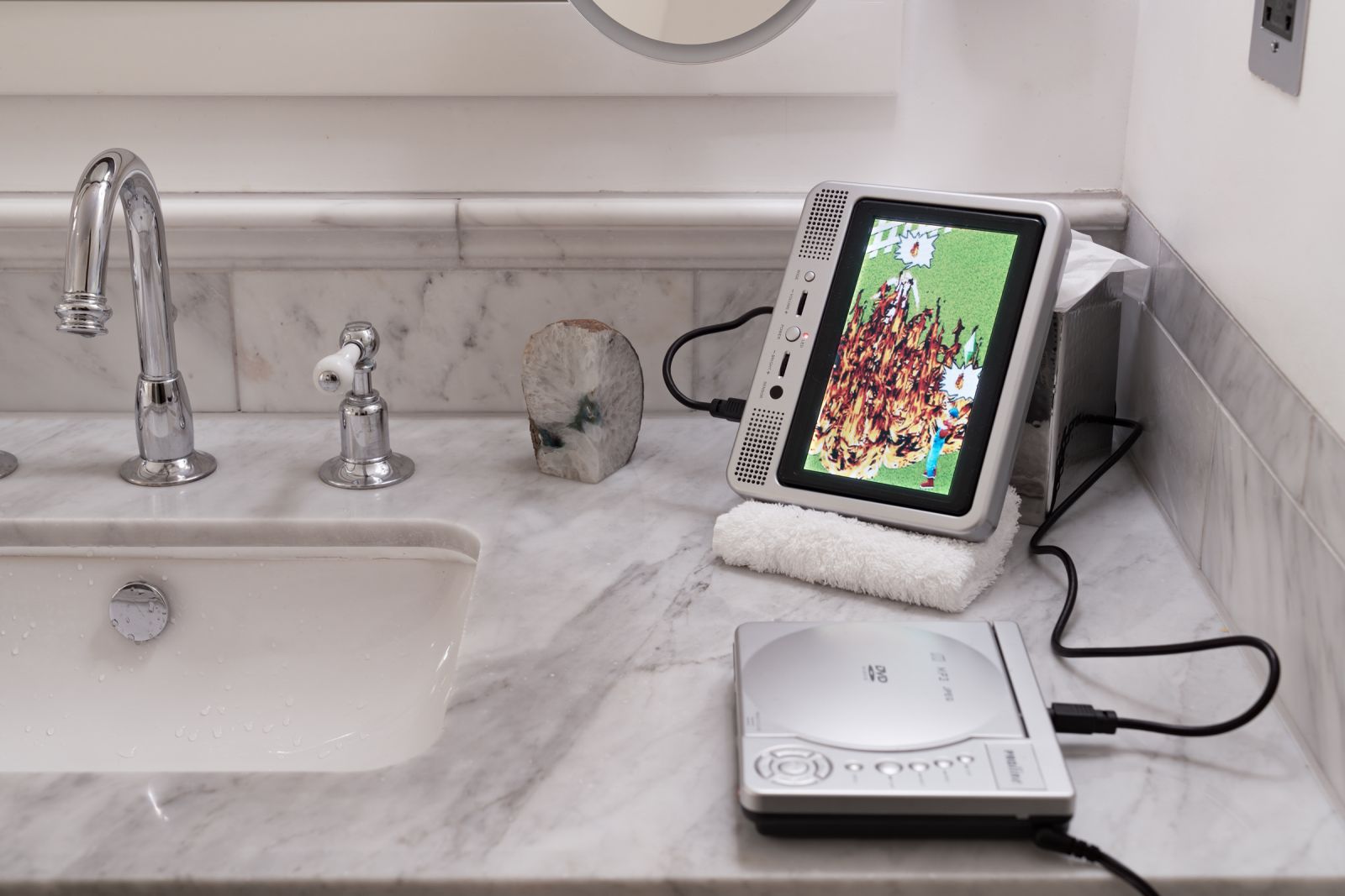 Portable media player on a marble vanity displaying a video game scene of characters burning. A sliced geode sits nearby.