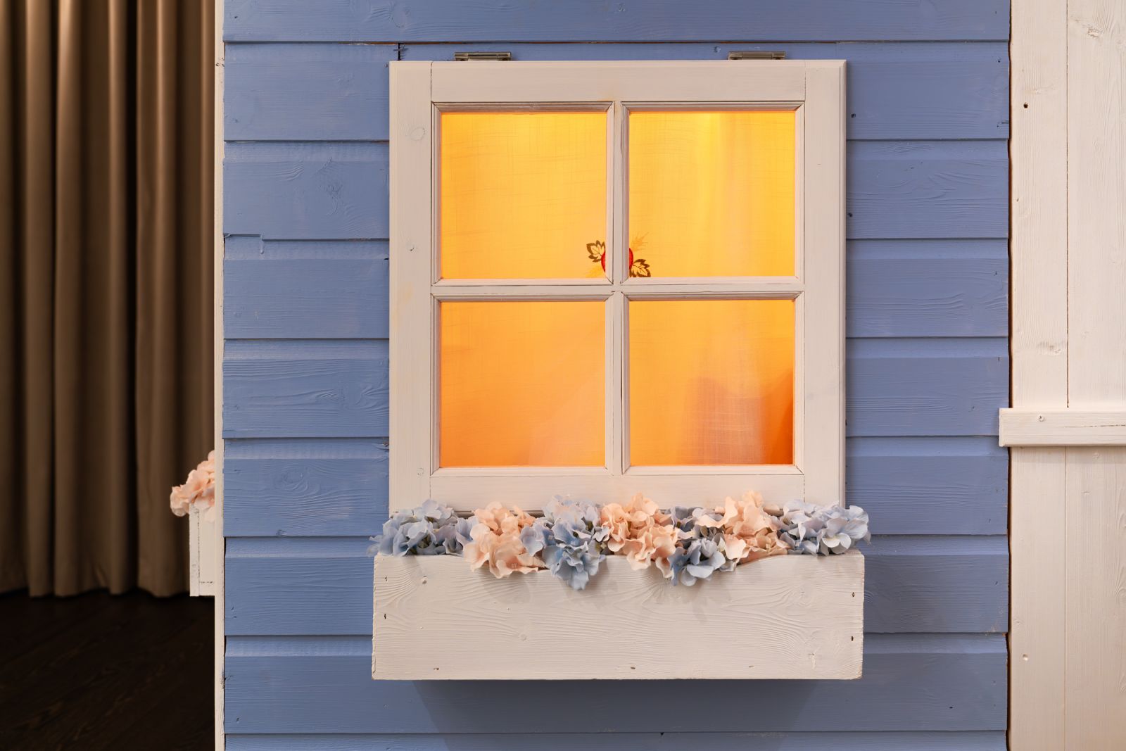 Close-up of a playhouse window with a white frame and glowing orange interior light. A flower box filled with artificial pink and blue hydrangeas sits below.