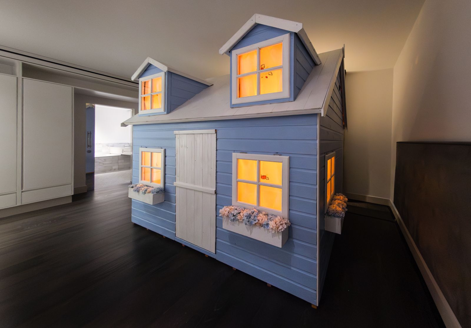 Full angle view of a two-story blue playhouse art installation indoors. Windows glow orange, contrasting with the dark gallery floor.