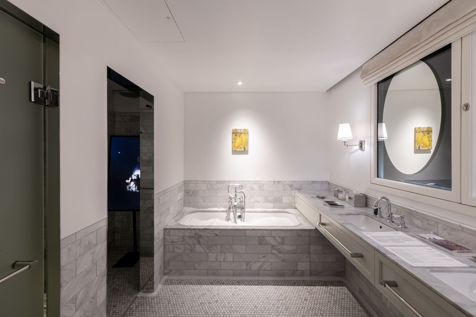 Wide shot of a bathroom installation showing a snow-filled tub, lemon painting, burning house video screen, and double vanity with a circular mirror.