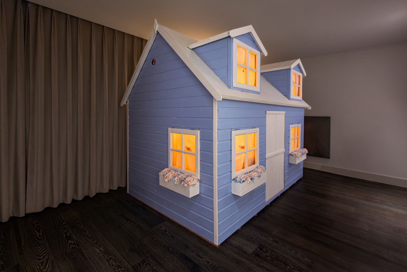 Large indoor art installation of a two-story blue playhouse with white trim. Windows glow with intense orange light suggesting fire, contrasting with pastel flower boxes.