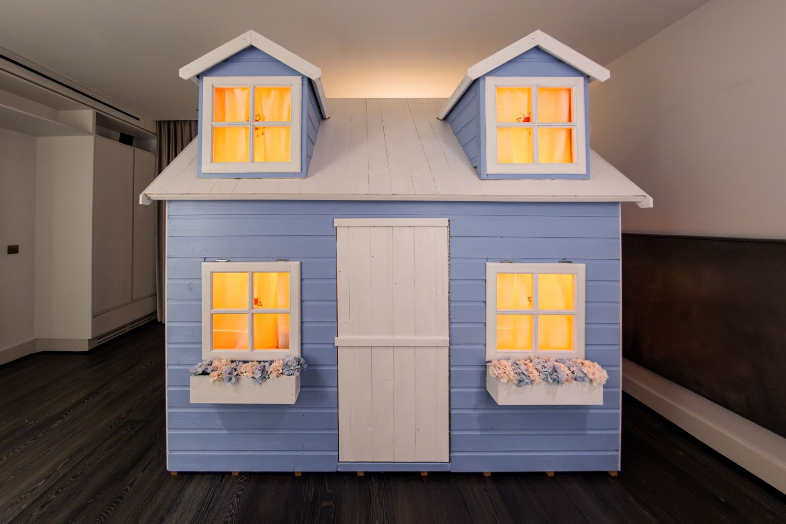 Front view of a symmetrical blue playhouse art installation. Four windows and dormers emit a flat, bright orange glow from within, flanking a central white door.