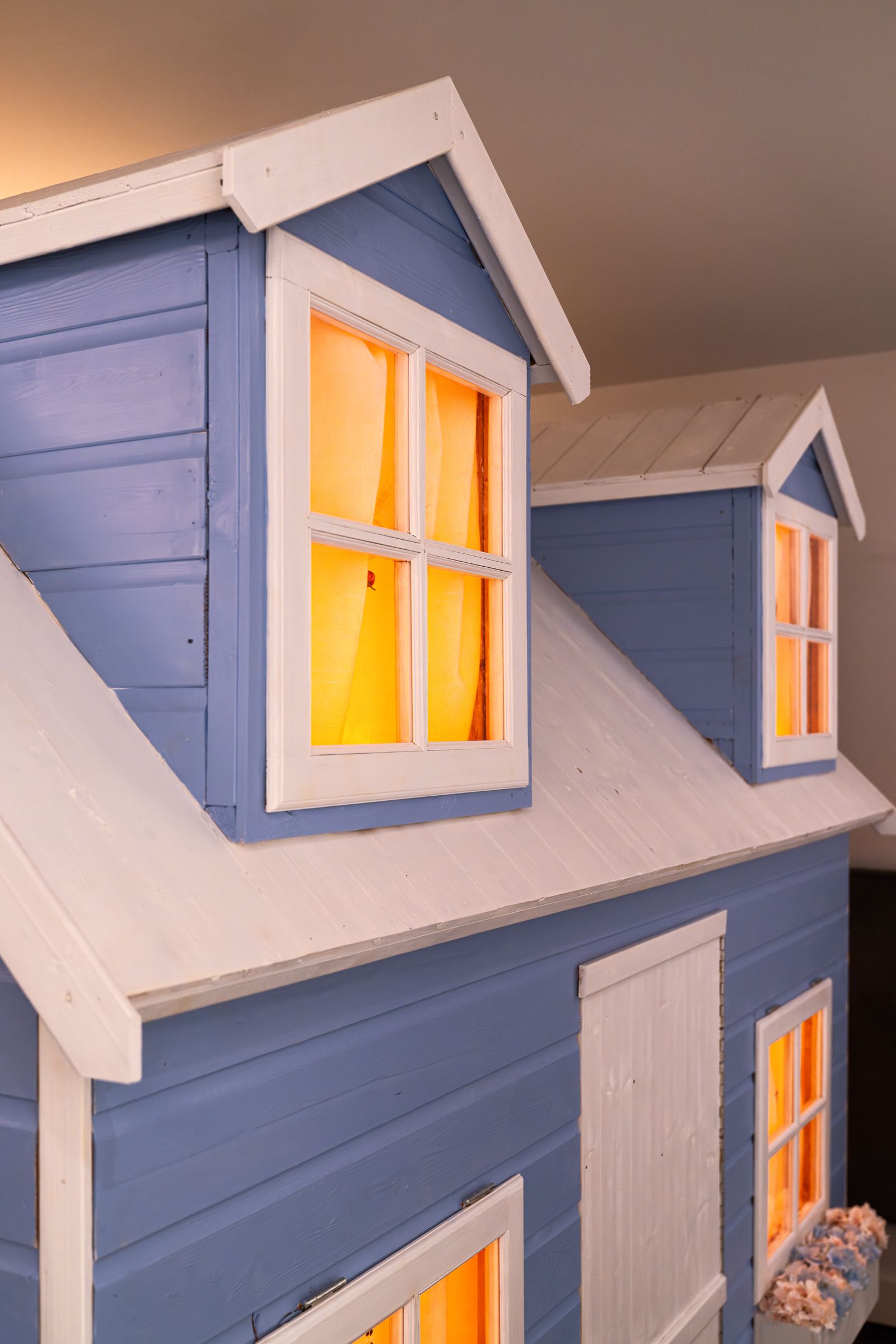 Angled detail of a playhouse roof showing painted blue wood texture and white roofing. Dormer windows radiate solid orange light.