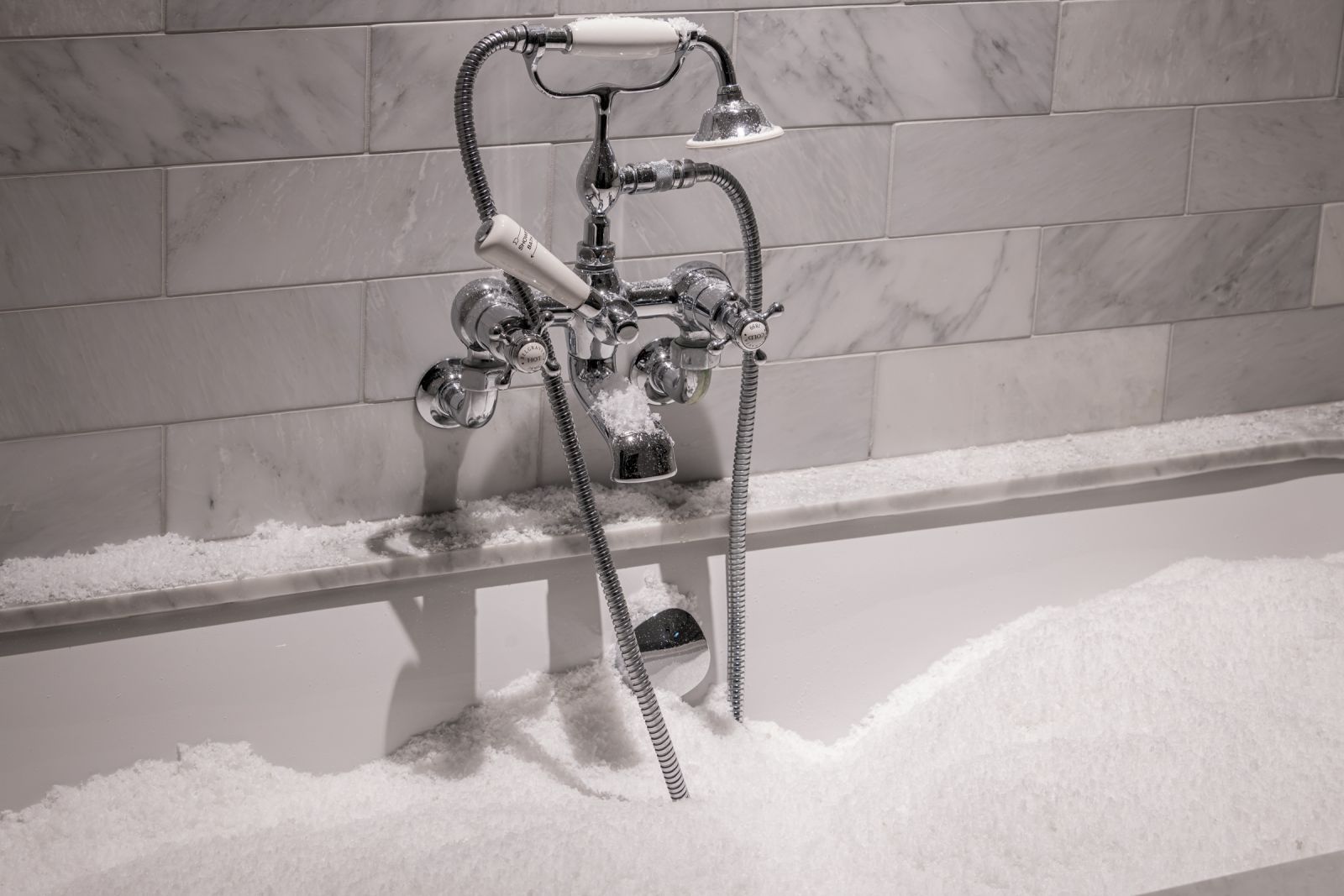 Detail of a chrome bathtub faucet and handheld shower head buried in white artificial snow, creating a frozen appearance.