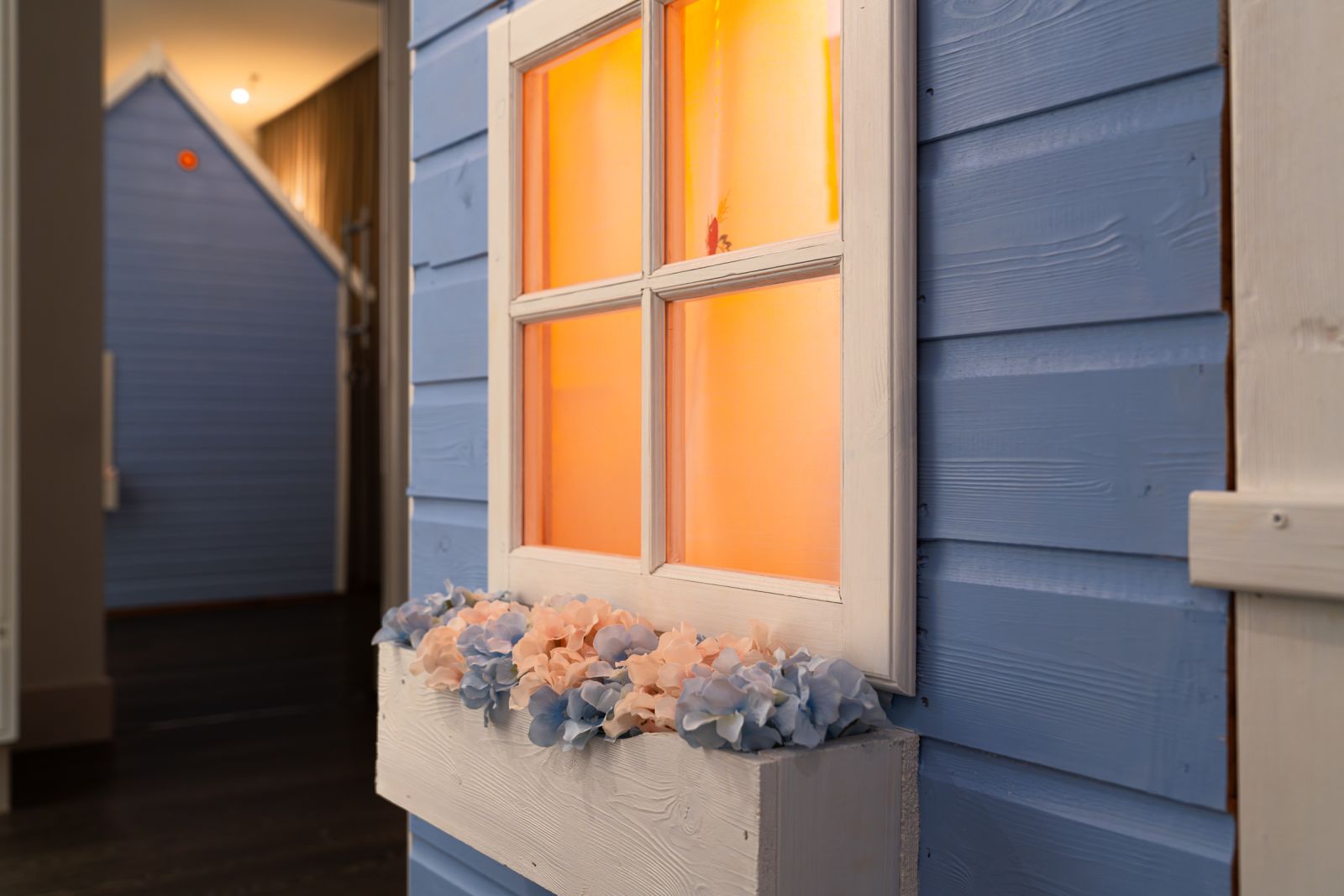 Detail of blue wood siding and white window frame illuminated by bright orange interior light, featuring a pastel flower box.