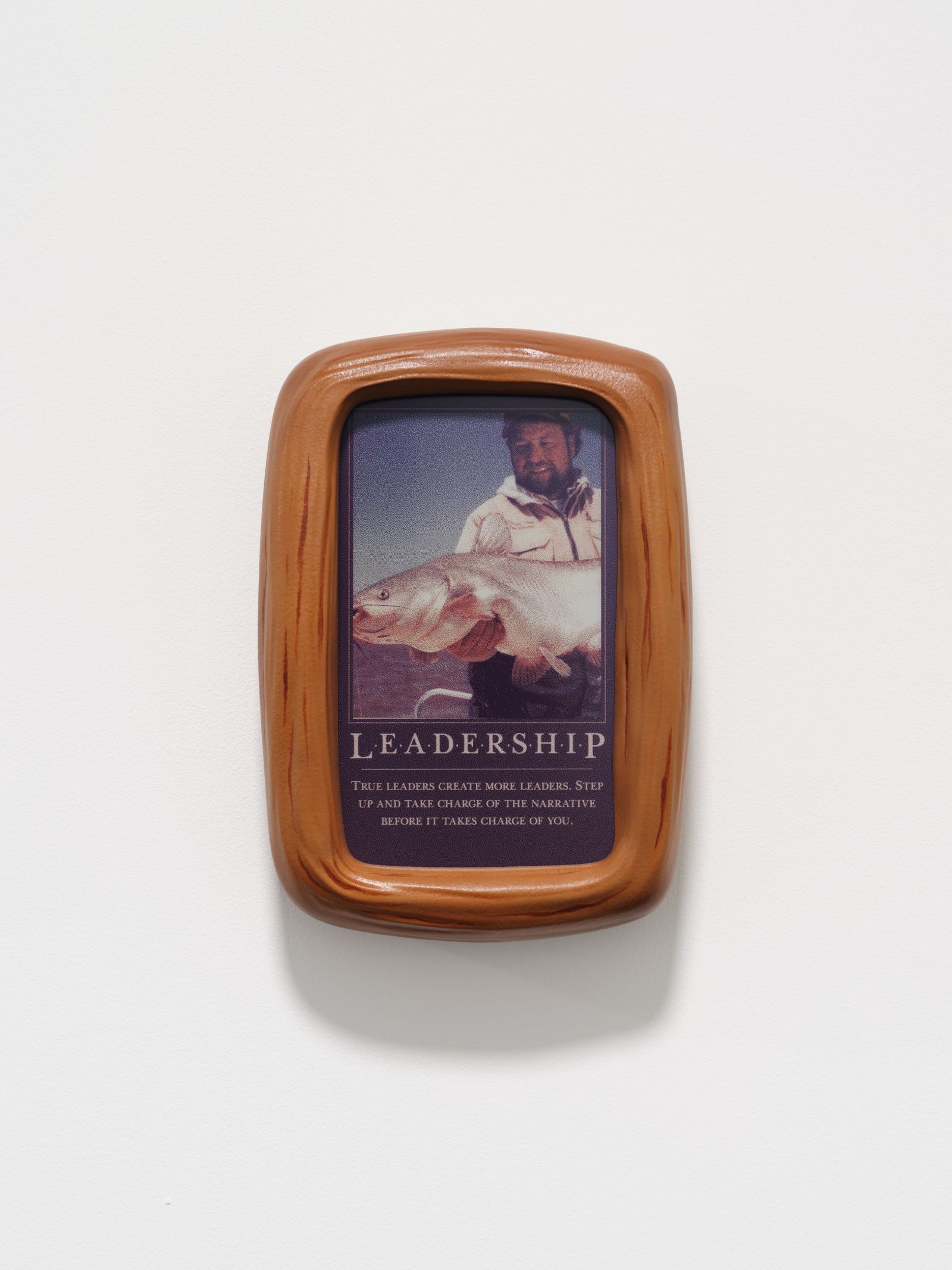 Close-up of a small brown frame on a white wall. The framed image features a man holding a large fish above the word LEADERSHIP and a short paragraph of text.