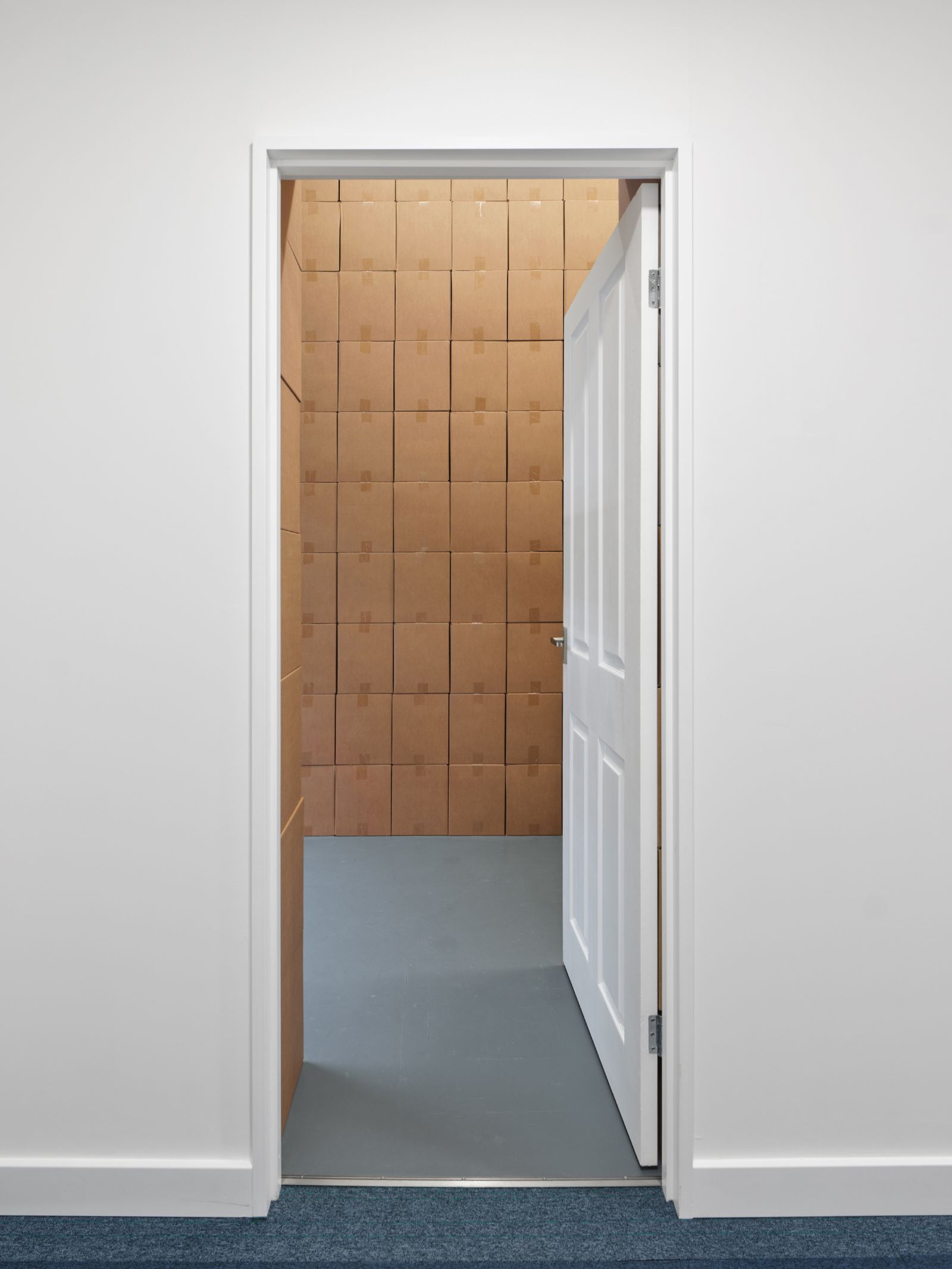 View from a white hallway with blue striped carpet looking straight through an open white door into a room constructed entirely of stacked cardboard boxes.