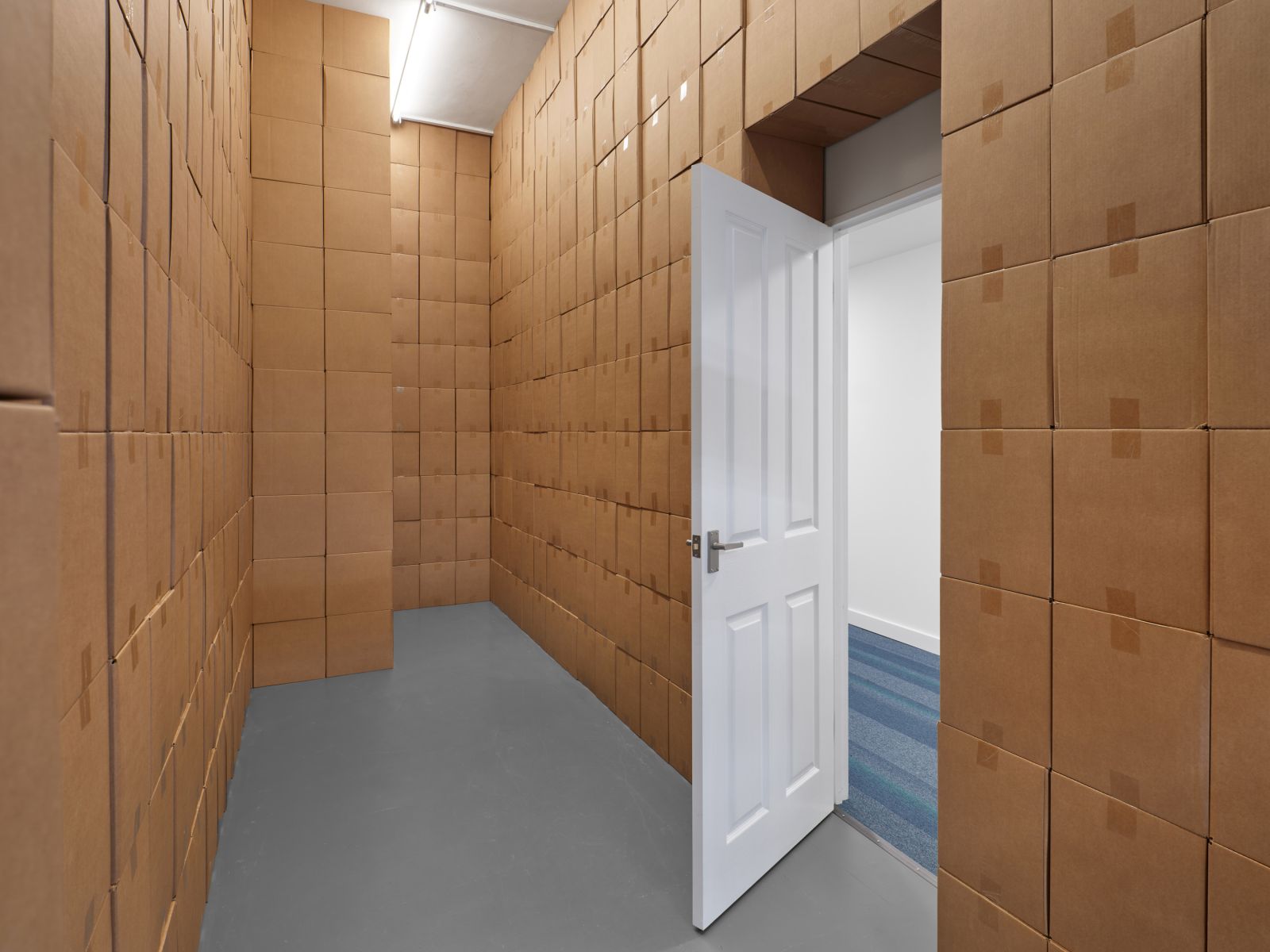 View from inside a room with walls built from uniformly stacked cardboard boxes, looking out through a partially open white door into an adjacent white hallway with a blue striped carpet.