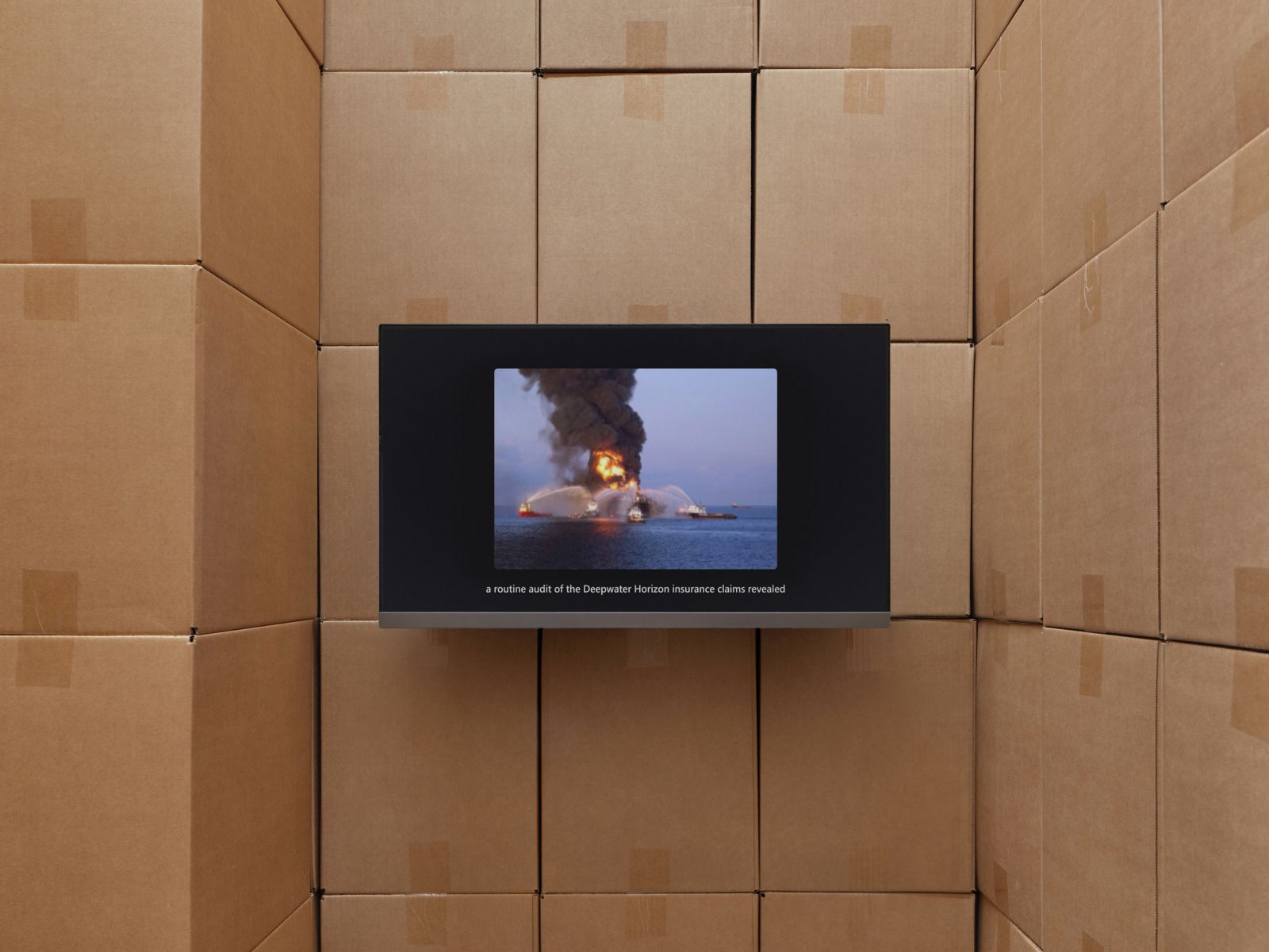 A flat screen embedded in a wall of cardboard boxes displaying video footage of an oil rig on fire at sea with subtitles about an insurance audit.