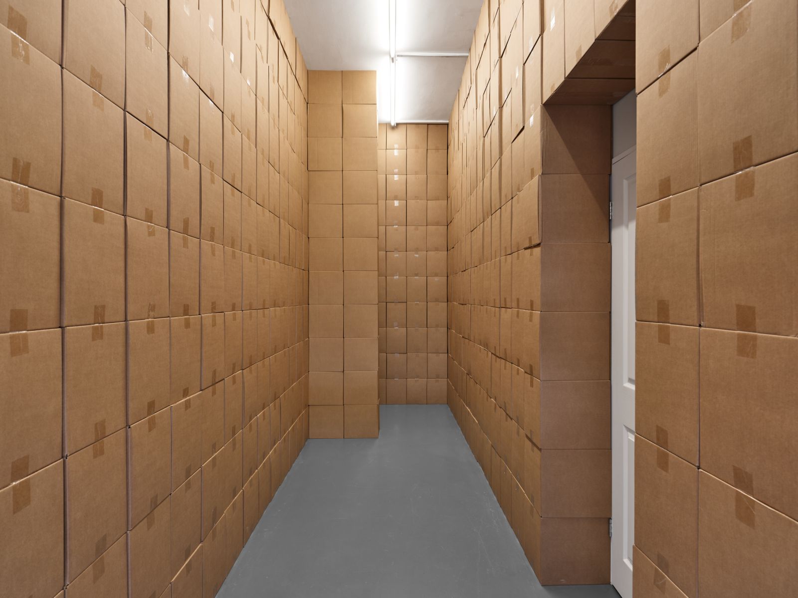 A narrow room with walls constructed entirely of uniformly stacked brown cardboard boxes, a grey floor, and a partially visible white door.