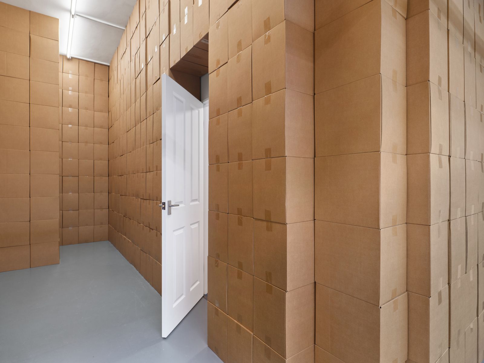 View of an exhibition space showing walls made of stacked cardboard boxes and a slightly open white door leading out of the room.