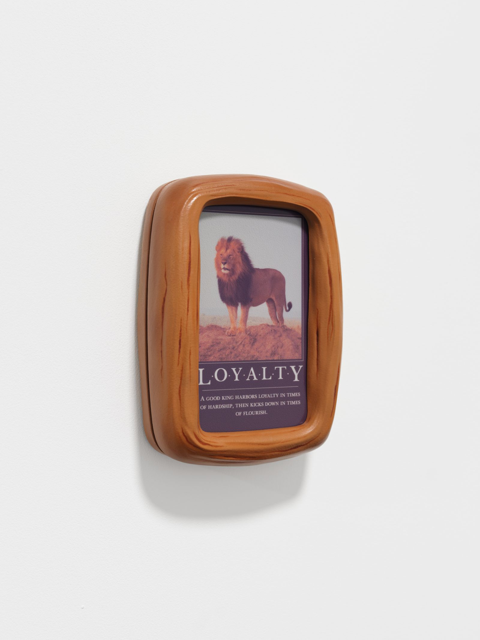 Close-up of a small brown frame mounted on a white wall. The framed image depicts a lion standing on dirt above the word LOYALTY and a short paragraph of text.