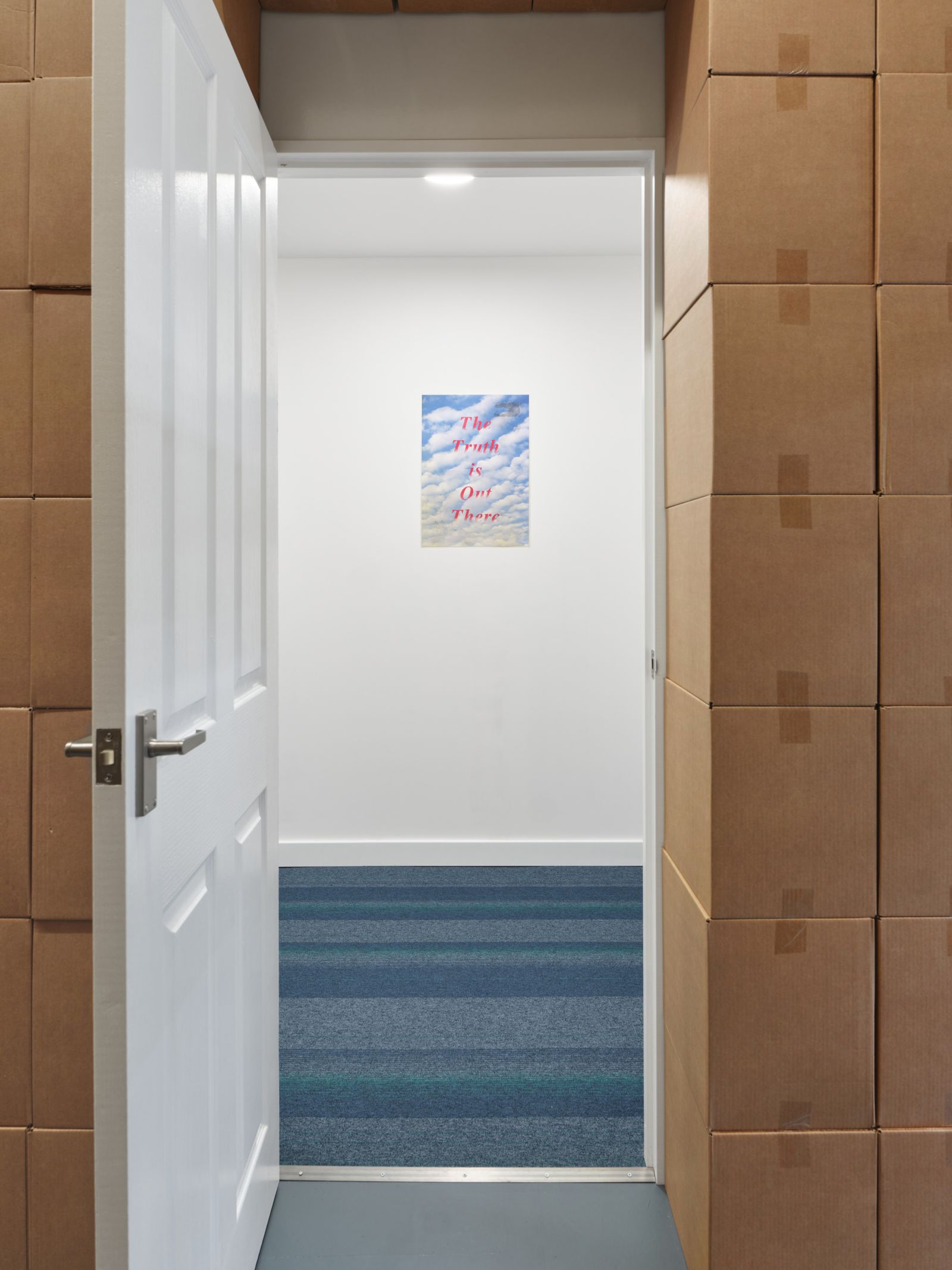 View looking through an open white door from a room with cardboard box walls into a white hallway with blue striped carpet and a poster on the far wall.