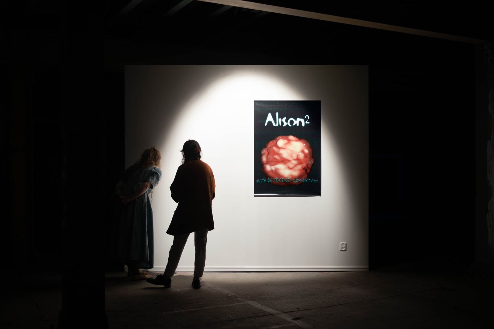 Two people standing in front of a white, highlighted wall in a dark industrial space