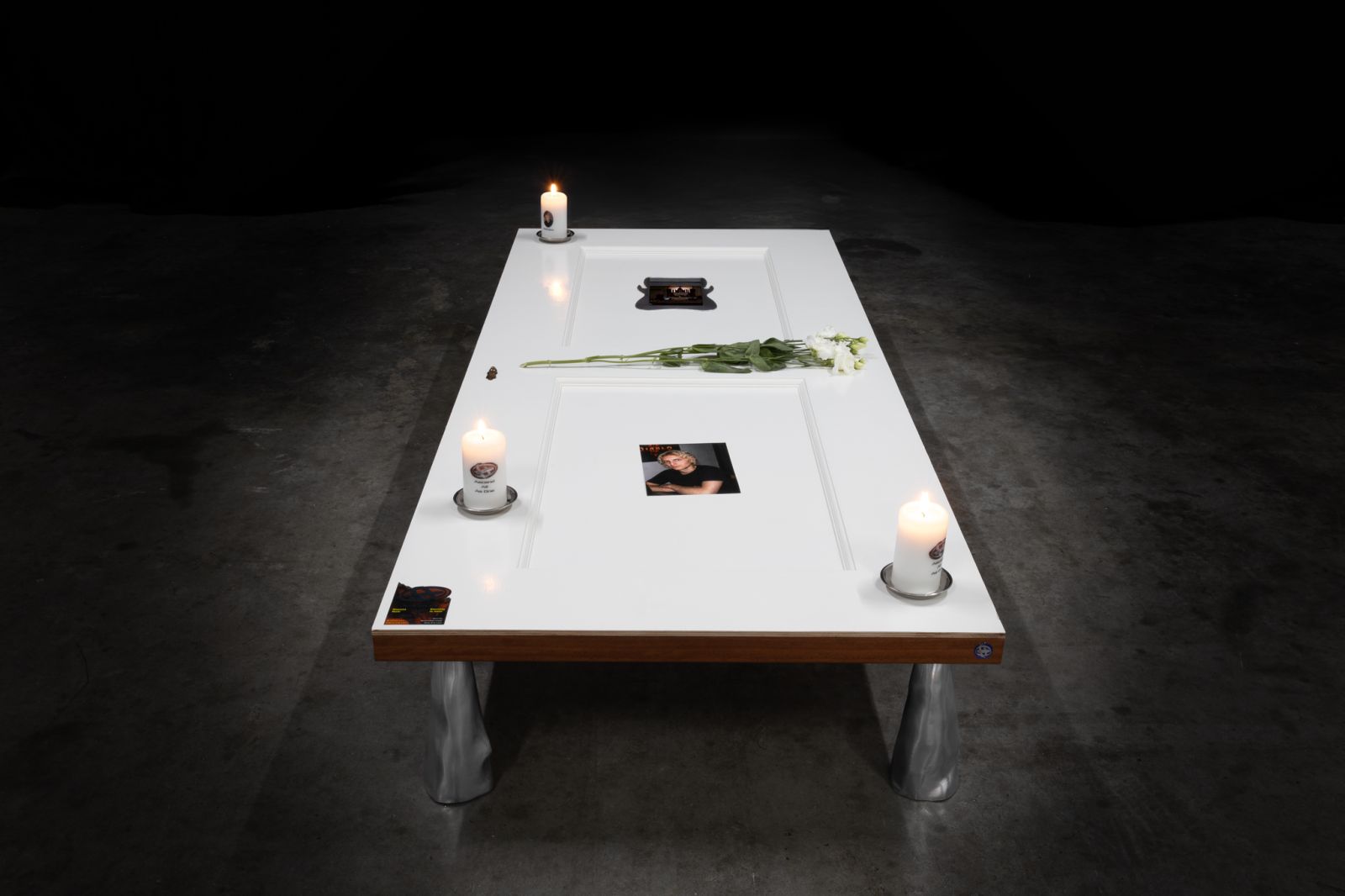 A white-painted door laying horizontally on 4 metal supports in a dark industrial space, on top of the door are 3 candles, flowers, a photo, stickers, and an embedded screen
