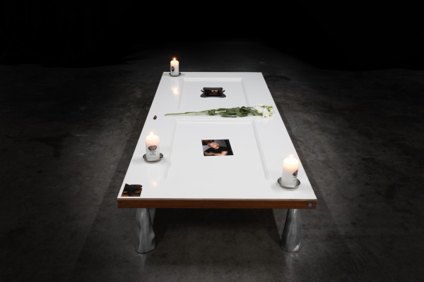 A white-painted door laying horizontally on 4 metal supports in a dark industrial space, on top of the door are 3 candles, flowers, a photo, stickers, and an embedded screen