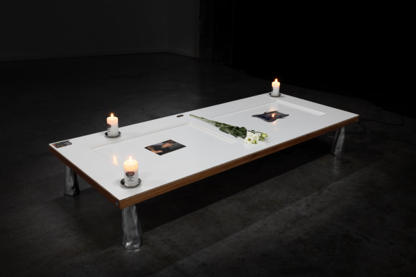 A white-painted door laying horizontally on 4 metal supports in a dark industrial space, on top of the door are 3 candles, flowers, a photo, stickers, and an embedded screen