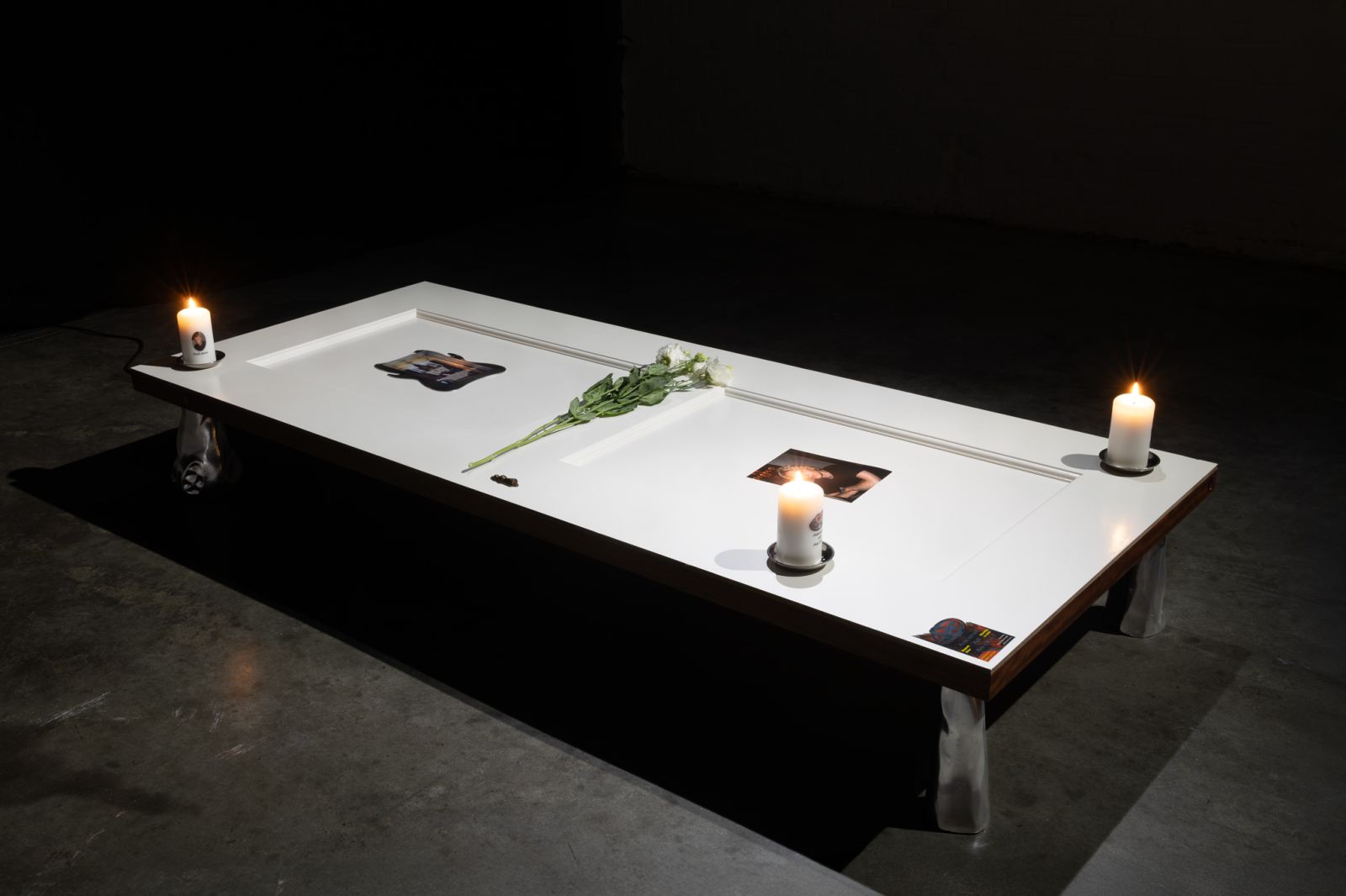 A white-painted door laying horizontally on 4 metal supports in a dark industrial space, on top of the door are 3 candles, flowers, a photo, stickers, and an embedded screen