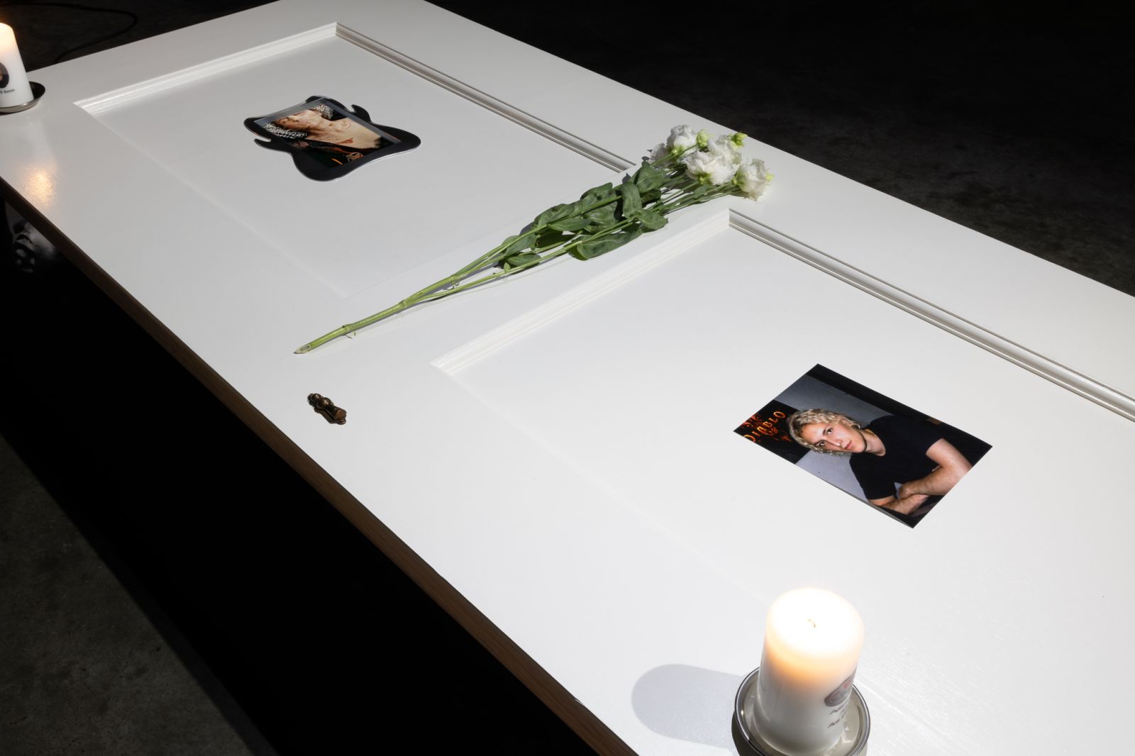 A white-painted door laying horizontally on 4 metal supports in a dark industrial space, on top of the door are 3 candles, flowers, a photo, stickers, and an embedded screen