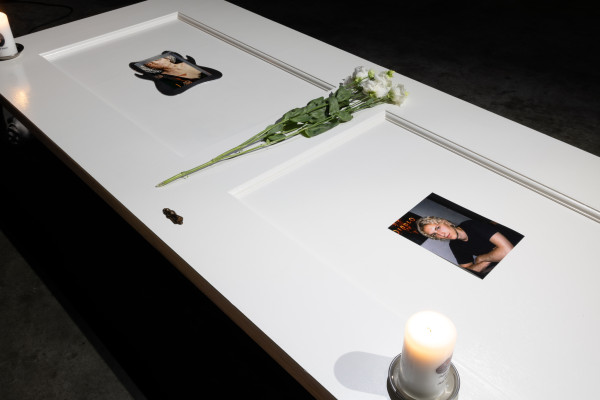 A white-painted door laying horizontally on 4 metal supports in a dark industrial space, on top of the door are 3 candles, flowers, a photo, stickers, and an embedded screen