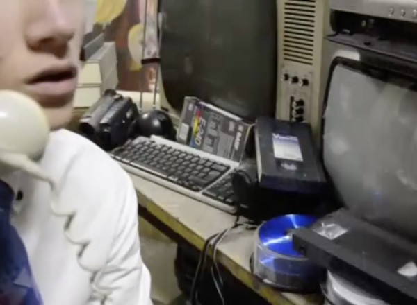 Man on the phone next to a messy desk with CRT monitors, computer and video equipment